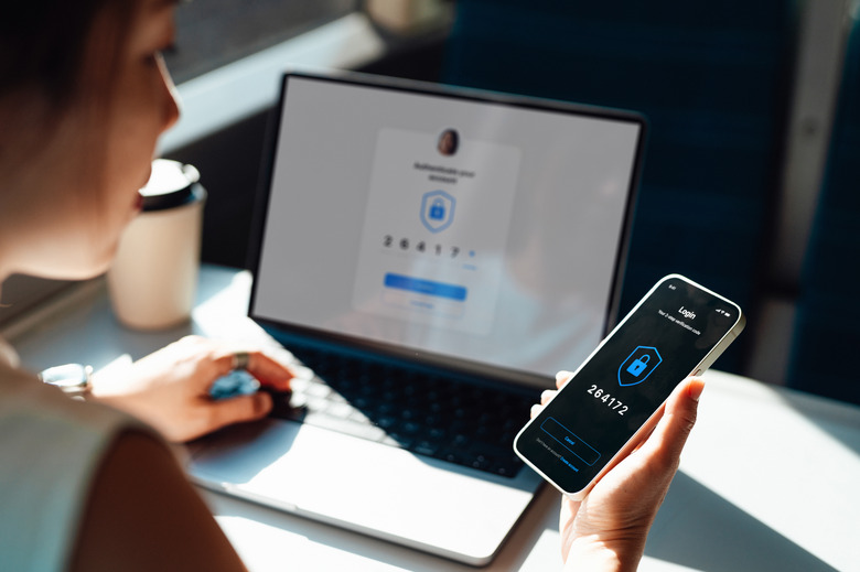 Over the shoulder view of young woman using smart phone while logging in personal banking account on laptop. Network security, data protection and privacy.