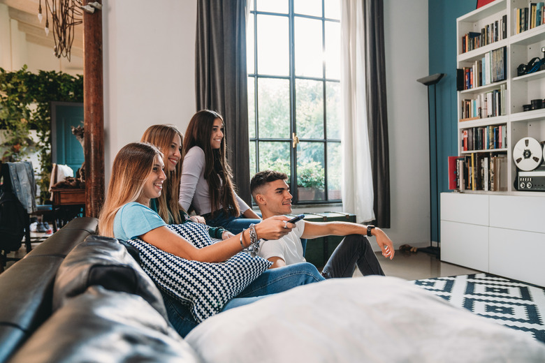 Four friends watching tv at home. They are sitting on the sofa, taking a break from homework.