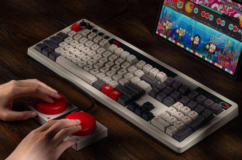 Product lifestyle photo of the 8BitDo Retro 108 Mechanical Keyboard. A person is mashing two big red buttons beneath the keyboard.