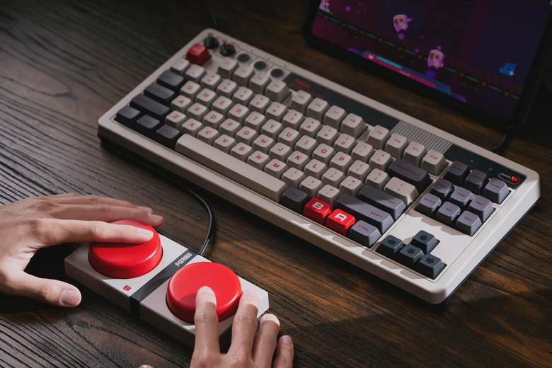 Product lifestyle photo of the 8BitDo Retro Mechanical Keyboard. The keyboard has Nintendo NES-inspired colors with deep (mechanical) keys. A pair of hands mash the two big red Super Buttons wired in front of it.
