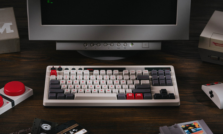 Product lifestyle image of the 8BitDo Retro Mechanical Keyboard. The mechanical keyboard has colors inspired by the NES console. 1980s gear surrounds it on a wooden desk.