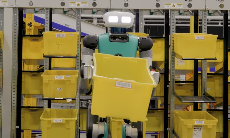 A humanoid robot with glowing eyes standing in a warehouse holding an empty yellow crate.