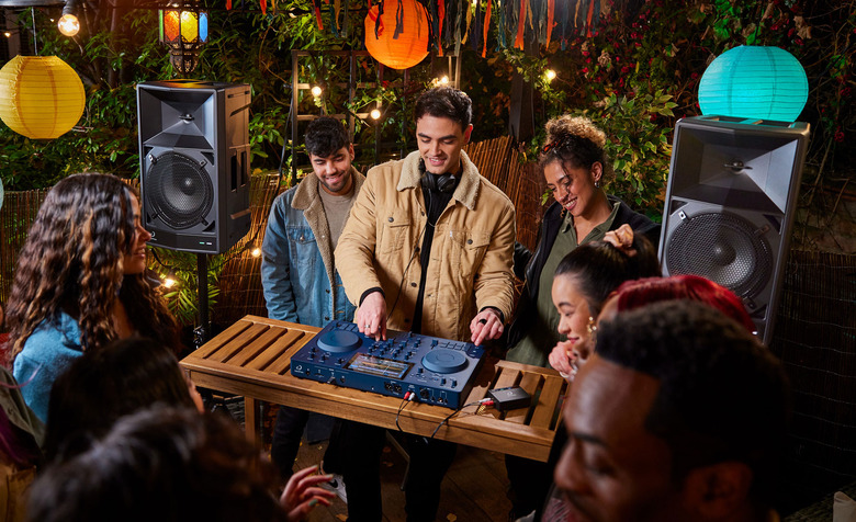 A DJ spins on an Omnis-Duo controller flanked by two Wave-Eight speakers amidst a small but lively party in a backyard.