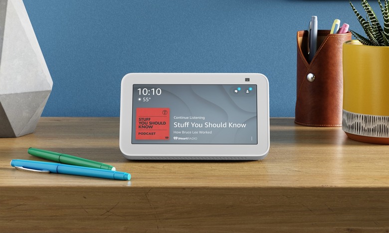A small smart display with a white border, the Amazon Echo Show 5, sits on a wood table next to various desktop accessories, displaying the time and the title of a currently playing podcast.