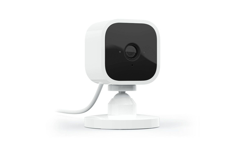 Marketing photo for the Blink Mini indoor security camera. The camera has a black front face and a white body. It’s facing the right with a power cord fading off to the left.