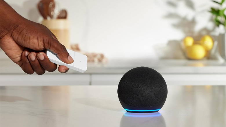 A hand holding the Amazon Basics Smart Remote, pointing it at an Echo speaker on a table.