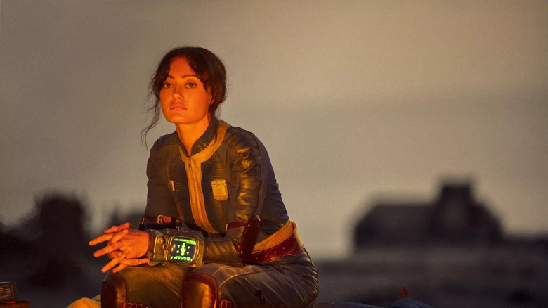 Ella Purnell (Lucy) in a still from the Fallout TV series. She sits, reflecting in front of a blurred desert landscape.