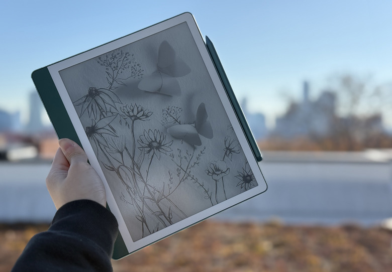 The Amazon Kindle Scribe 2nd gen being held up in mid air, with a Premium Pen attached to its right side. In the background is the NYC skyline.