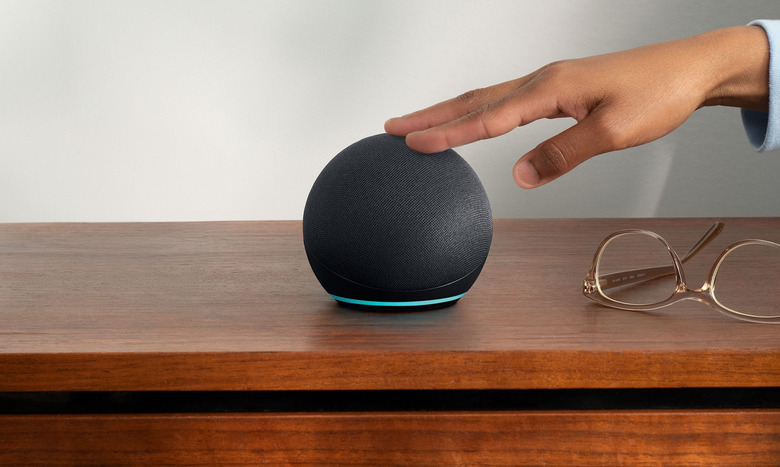 Amazon's Echo Dot speaker sits on a wooden nightstand. A person's hand is reaching out to touch the top of the speaker. Probably to snooze the alarm. 
