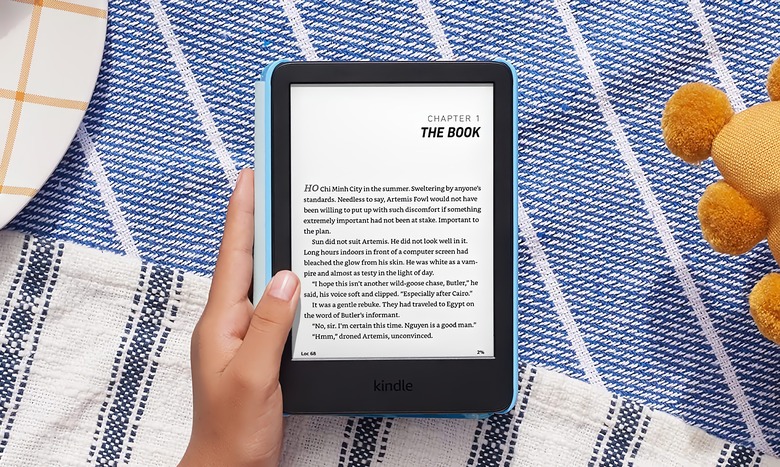 Amazon marketing photo of the Kindle Kids e-reader. A child's hand holds the device (showing a page from a book) with a colorful blanket and edge of a plush toy behind it.