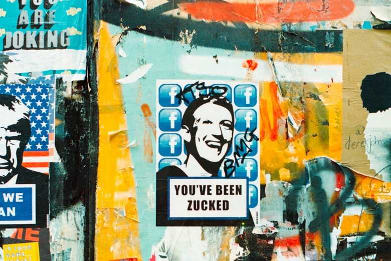 Photo of an urban wall, featuring stickers. Prominently centered is one of Mark Zuckerberg smiling, with the caption, 