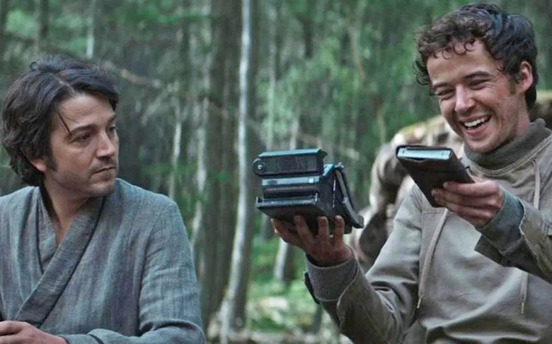A still image from the tv series 'Andor' showing the main character sitting next to someone who's holding a device that appears to look like a folding polaroid camera. 