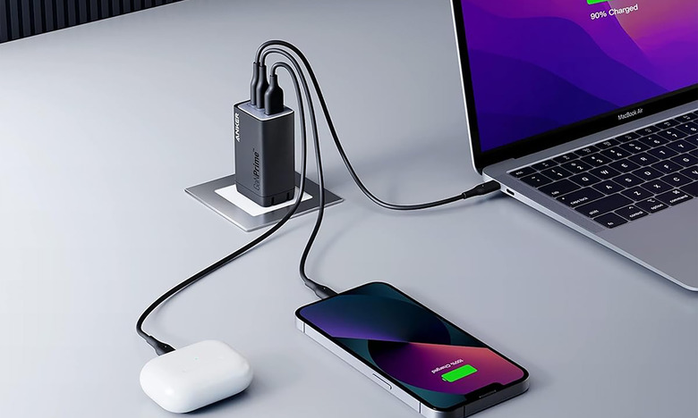 The Anker GaNPrime 65W wall charger is plugged in and charging up a laptop, smartphone and AirPods on a gray desk. 