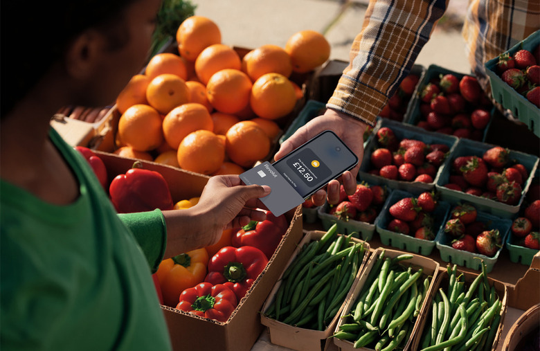 A person wearing a green shirt holding a credit card over an iPhone to pay for their purchase. Oranges, bell peppers, strawberries and string beans in containers are in the  background.