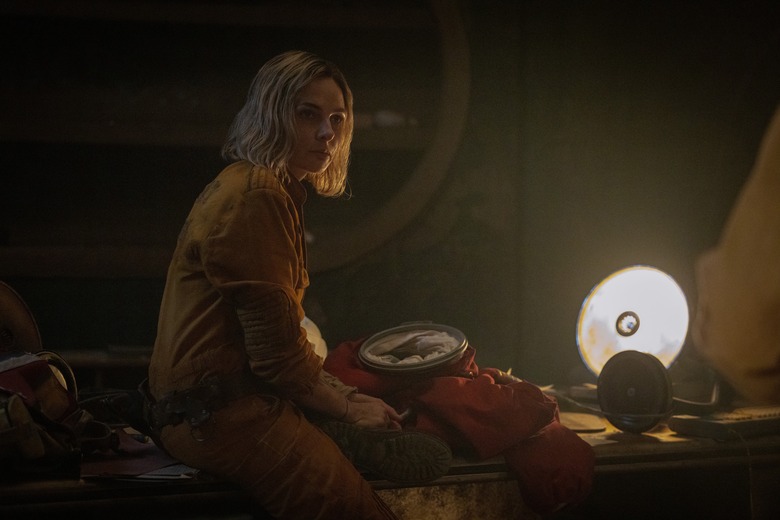 A woman wears a yellow jumpsuit and sits on a bed while a lamp illuminates her from behind.