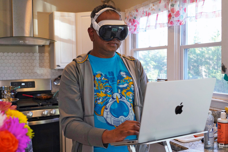 A man stands in front of his laptop in a kitchen while wearing the Apple Vision Pro AR/VR headset.
