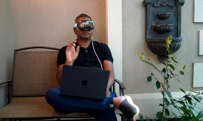 Engadget editor Devindra Hardawar is pictured sitting on an outdoor bench wearing the Apple Vision Pro headset and holding his hand up in a gesture in front of him