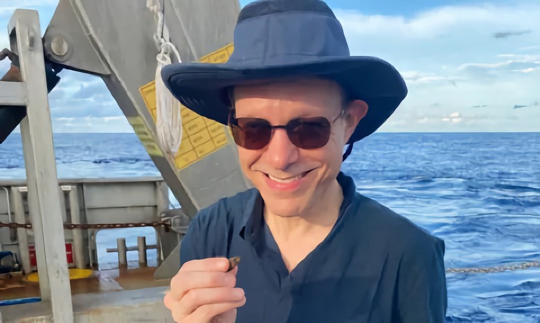 Photo of Harvard astrophysicist, on a boat with sunglasses and hat, holding a small object in his hand.