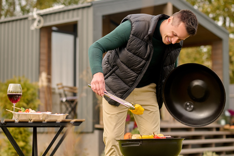 Smiling man leaning over the open brazier and gripping the slice of bell pepper with barbecue tongs