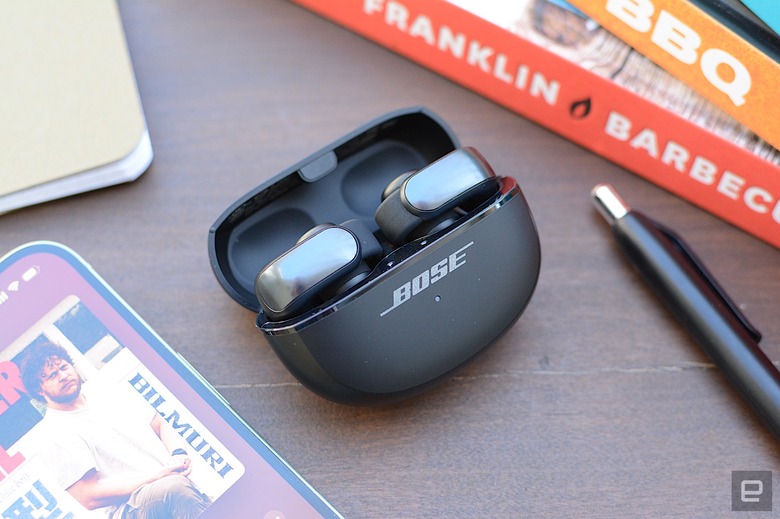 A pair of Bose Ultra Open Earbuds both sit in their charging case with the lid open on a wooden desktop surrounded by books, a smartphone and a pen.