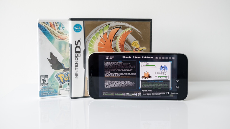 An iPhone 12 sits in front of the cases for Pokemon Heart Gold and Pokemon X. It displays the Claude Plays Pokemon Twitch stream. 