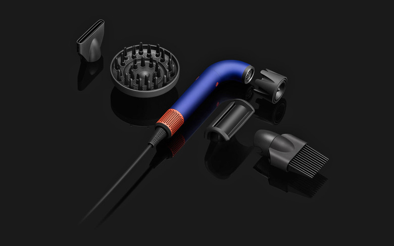 The components of the Dyson Supersonic R hairdryer are laid out next to each other on a black surface. in the middle is the main J-shaped tubular dryer body with its several attachments.