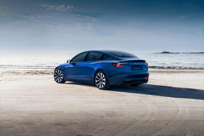 A blue Tesla sedan on a beach. Tire tracks in the sand.