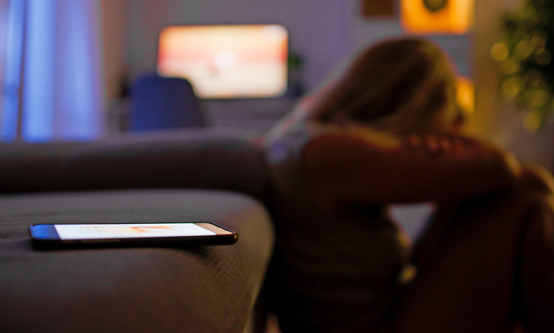 A seemingly depressed woman sits on floor in front of a couch hugging her knews, with a mobile phone in the foreground on the sofa.