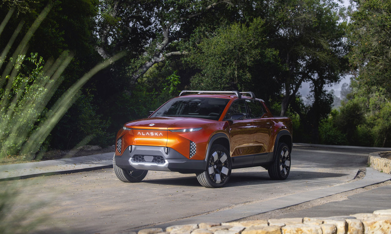 Image of an orange Fisker Alaska flatbed EV driving down a quiet road in front of some dark green foliage.
