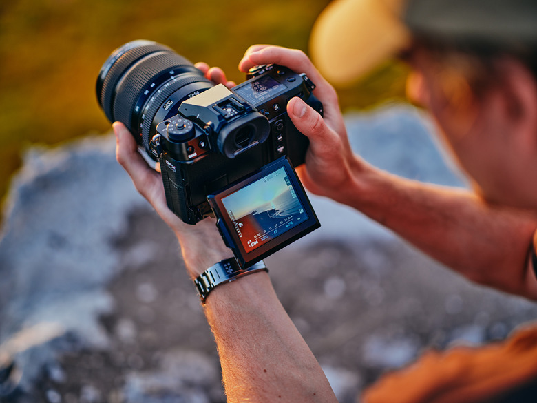 A person holding the Fujifilm GFX 100S II camera with its viewfinder folded out. View from slightly above their left shoulder.