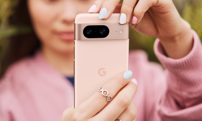 A woman in a pink sweater holds up a pink smartphone, the Google Pixel 8, in front of the camera, showing its back and camera system.