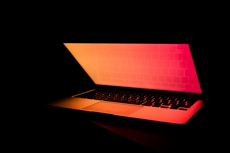 A Macbook laptop pictured in a dark setting half opened with an orange and red glow coming from the screen 