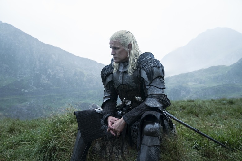 Still from House of the Dragon. A man with long white hair wears a suit of armor as he sits on a mound of grass. There are mountains in the background.