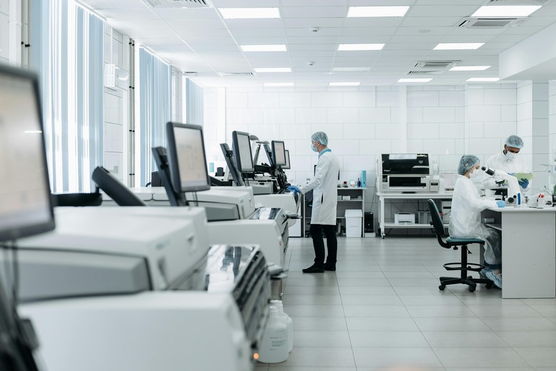 A white room with multiple standing computer monitors, with a doctor in a lab coat and hair net in front of one. Two other doctors sit behind, one looking through a microscope