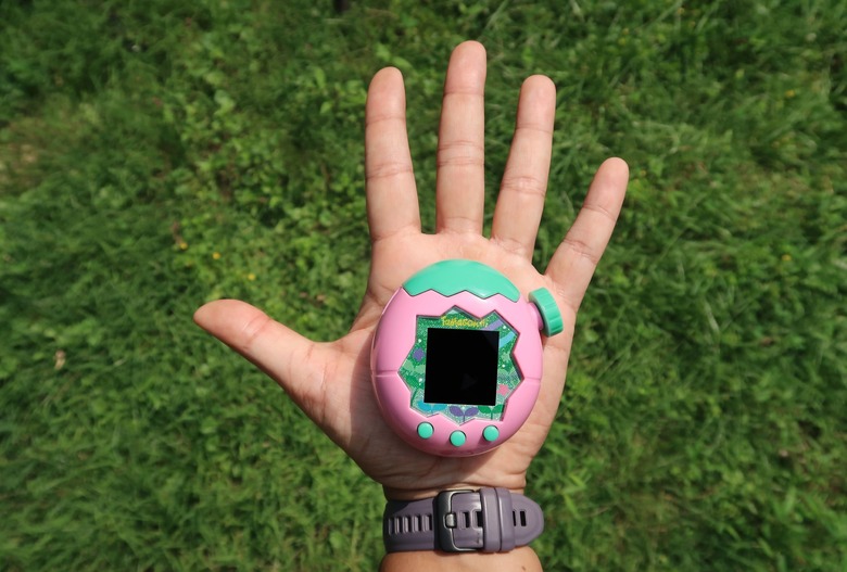 A pink and green Tamagotchi Paradise (Land model) pictured in the palm of an outstretched left hand, with green grass in the background