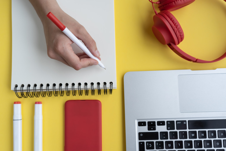 A high angle shot of a person taking notes in a notebook with headphones, pens and a laptop on a yellow surface