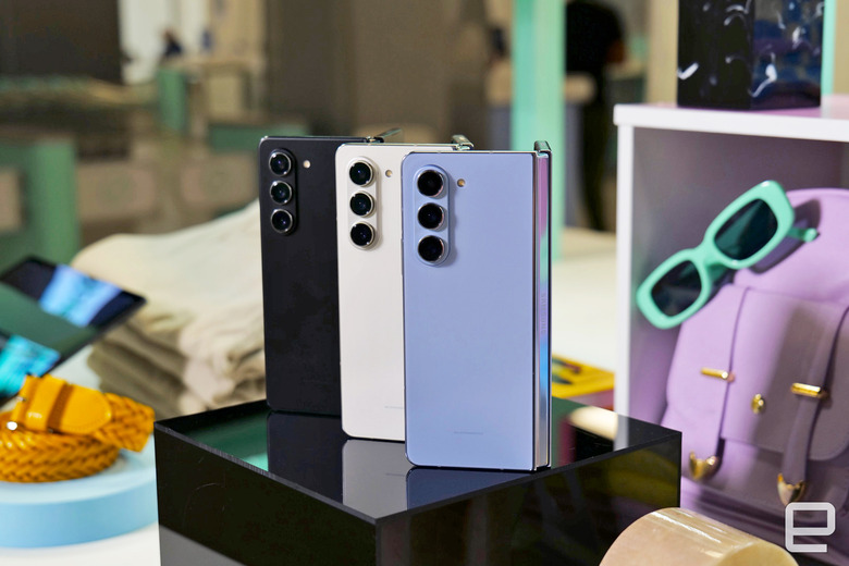 Three of the Samsung Galaxy Z Fold 5 phones stand on end open 45-degrees and showing the exterior of the phone, while sitting amongst a variety of pastel colored props, bags, sunglasses and folded clothing.