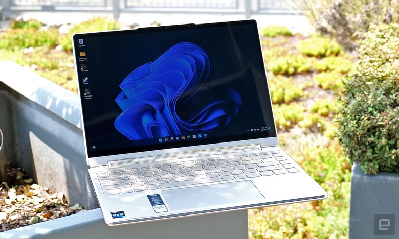 The Lenovo Yoga 9i laptop in white sits open and facing the camera as it sits on the edge of an outdoor planter, with lots of plants and bushes in the background.