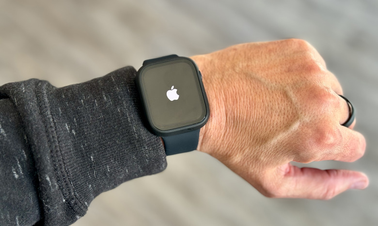 A left arm / wrist / hand wearing an Apple Watch with a white Apple logo on its otherwise black screen. Blurred gray background.