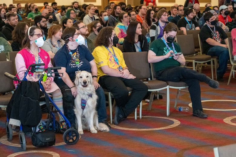 A room full of audience members at a Games Done Quick charity speed-running event. In the front row, someone holds a cream-colored golden retriever, who smiles for the camera.