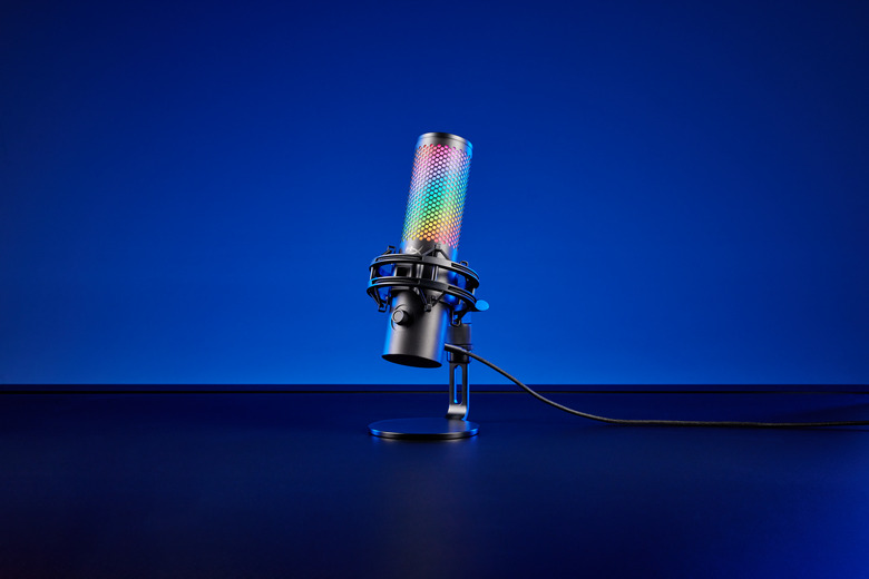 Product image of the HyperX QuadCast 2 S gaming mic. The mic (with over 100 LEDs) sits on a black desk in front of a blue background. Dramatic shadows.