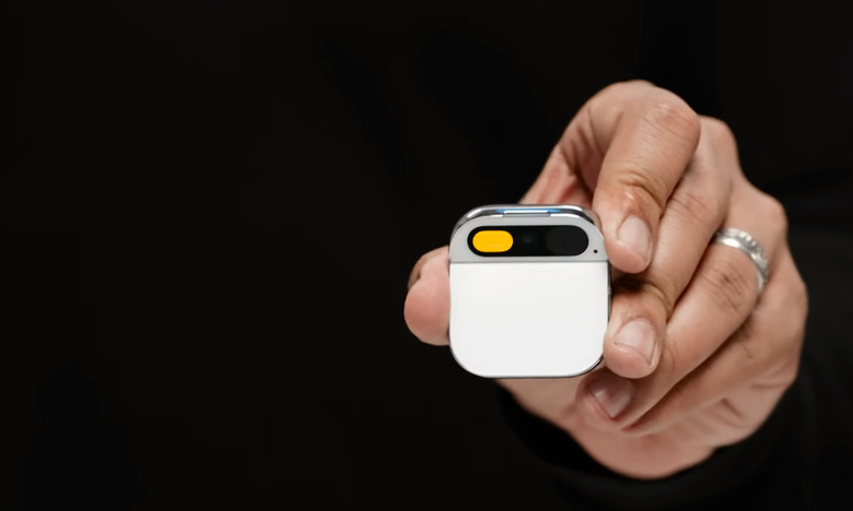 The hand of co-founder Imran Chaudhri holding the Humane AI Pin. Closeup. The small device has a white face, and is square with round corners and a section at the top for a camera.