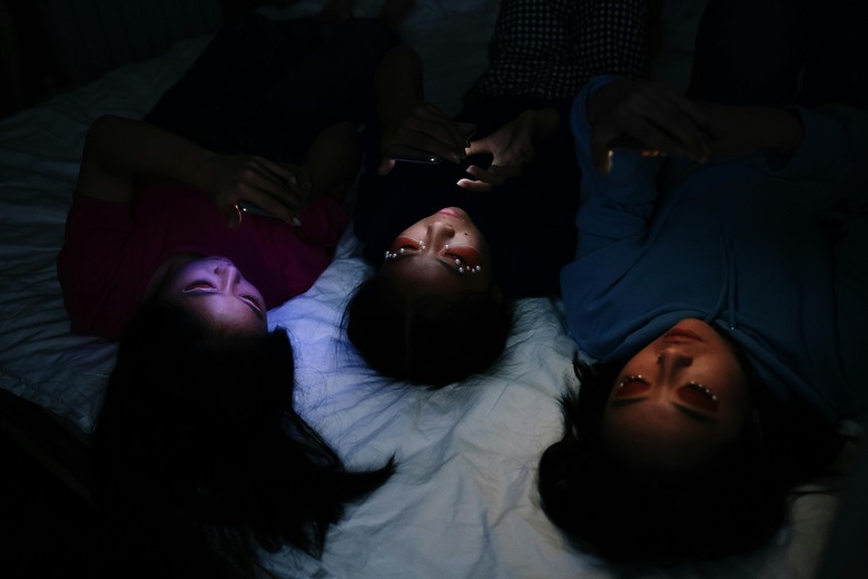 Three young people lay on their backs in bed in the dark, with their faces lit by the glow of their phones