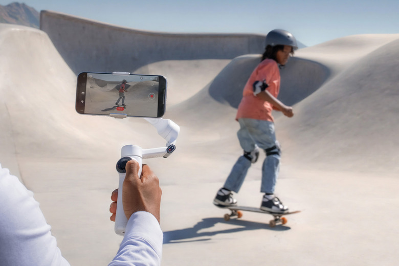 A person holding a smartphone on a Flow 2 gimbal. They're filming a child skateboarding in a skate park.