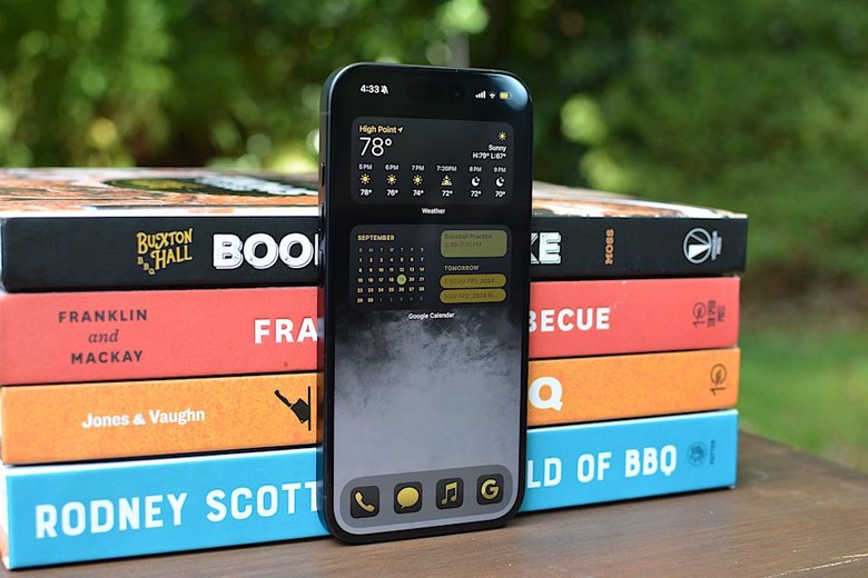 An unlocked iPhone 16 leaning against a stack of books outside.
