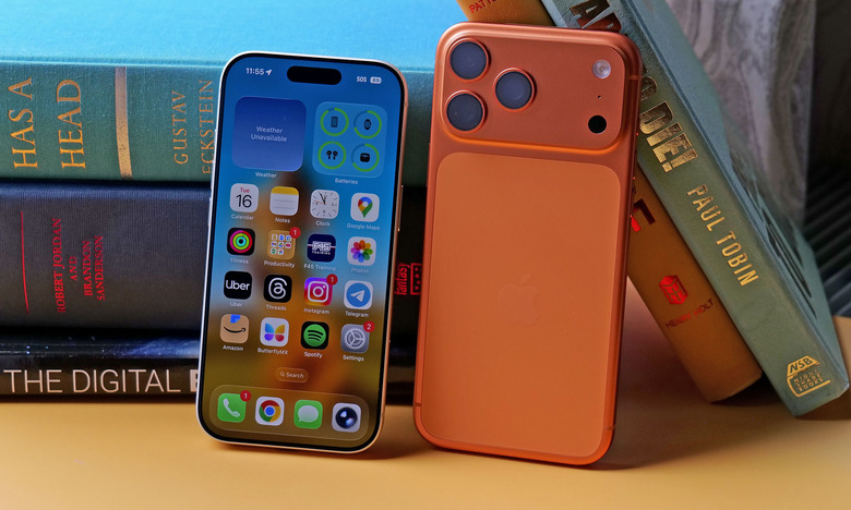 An iPhone 17 Pro with its screen facing out and an iPhone 17 Pro Max with its rear facing out. Both are leaning against a pile of books.