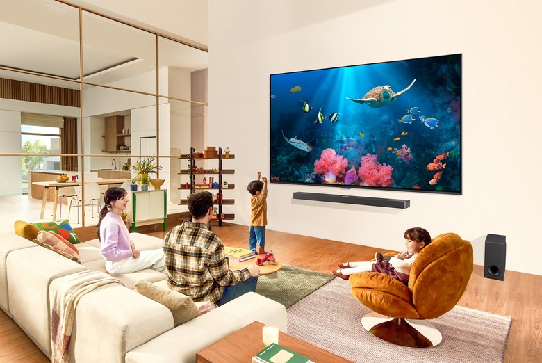 LG's 2024 TV lineup promotional image showing a family in a modern living room in front of a large wall-mounted 98-inch QNED TV showing an underwater scene with flsh.