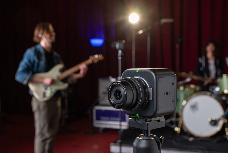 Lifestyle marketing image of the Logitech Mevo Core livestreaming camera. It sits on a tripod with a rock band playing in a studio behind them.