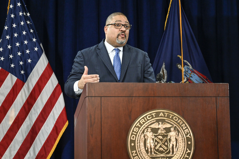 A person in a suit behind a wooden podium with the American flag in the background.