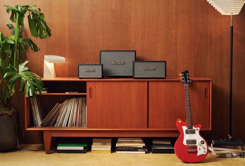 A living room media console has three Marshall speakers on it, with records in the cabinet and a guitar leaning against it.